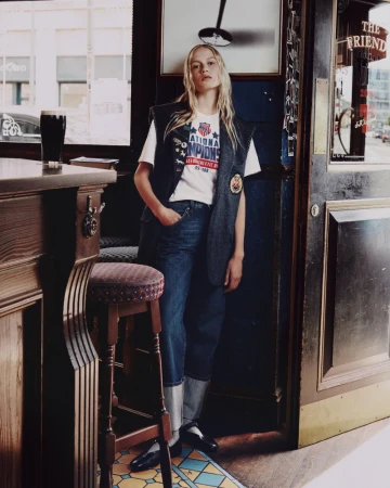 Model standing inside a pub wearing a white graphic t-shirt, dark vest, and cuffed denim jeans