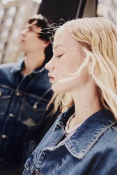 Close-up portrait of a blonde woman wearing a denim jacket and pearl necklace with a man in the background outdoors.