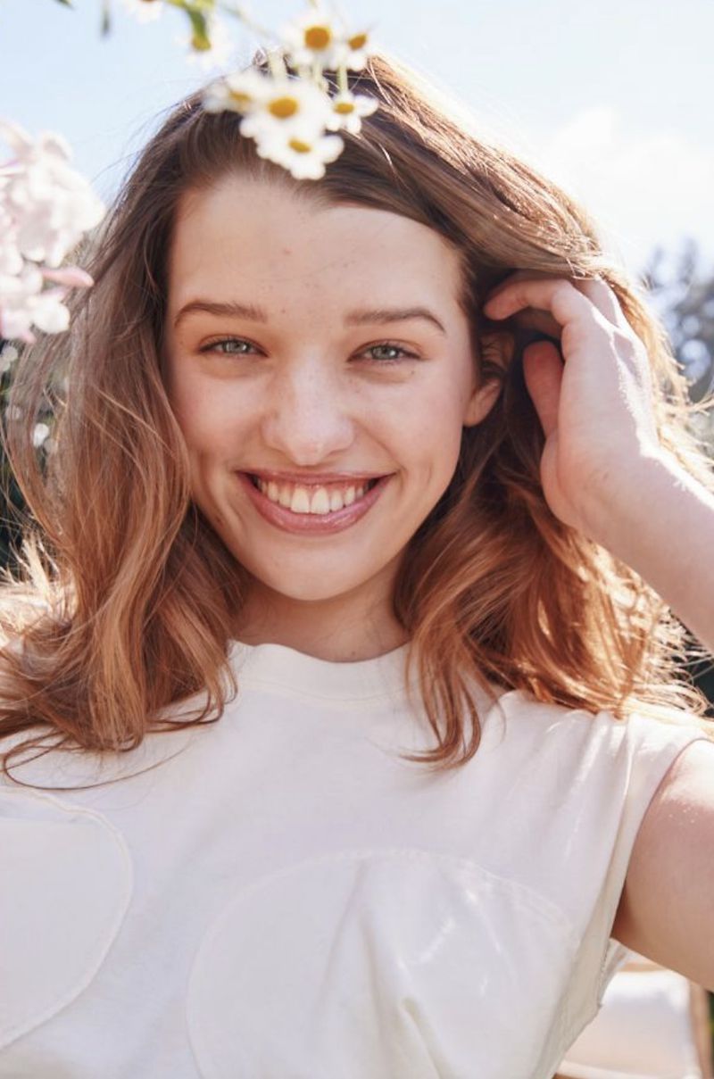Smiling woman with shoulder-length hair, wearing a white top, surrounded by white and yellow flowers in an outdoor setting.