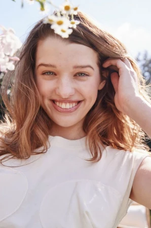 Smiling woman with shoulder-length hair, wearing a white top, surrounded by white and yellow flowers in an outdoor setting.