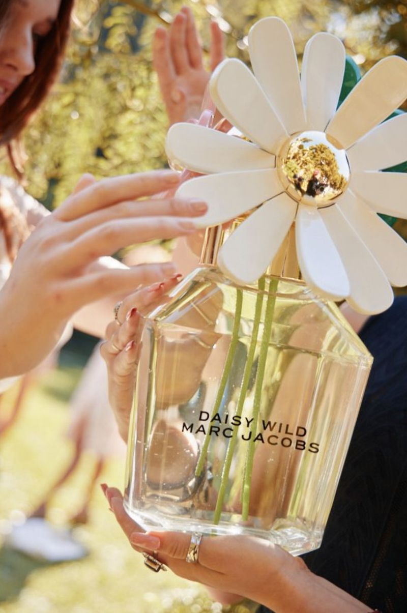 Daisy Wild Marc Jacobs perfume bottle held by two people in a bright, outdoor setting with blurred green background, flower bottle cap.