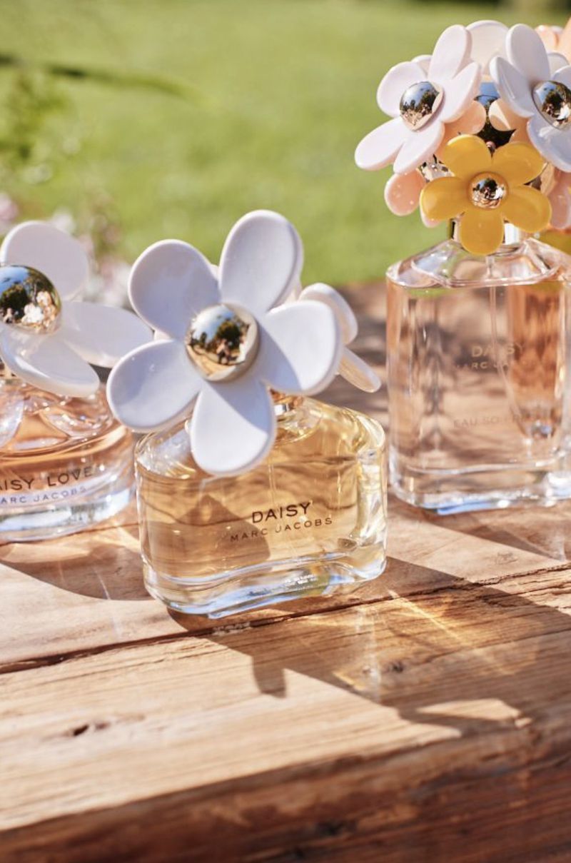 Three Marc Jacobs Daisy perfume bottles with distinctive flower caps resting on a rustic wooden surface outdoors.