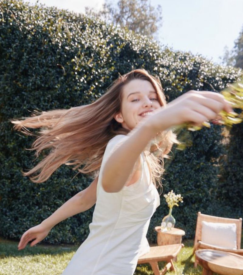A young woman in a white dress twirls happily on a grassy lawn in bright sunlight, with a backdrop of lush green bushes.