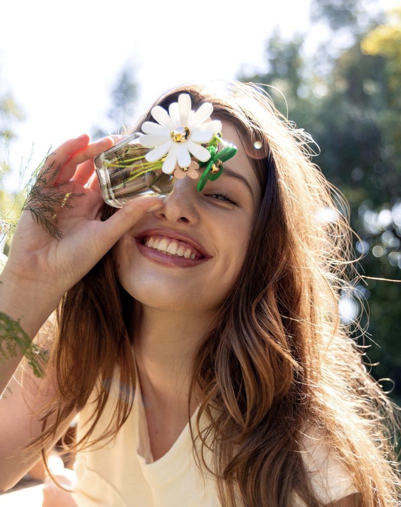 Smiling woman with long hair holds up a clear glass jar with flowers to her eye in a sunlit outdoor setting.