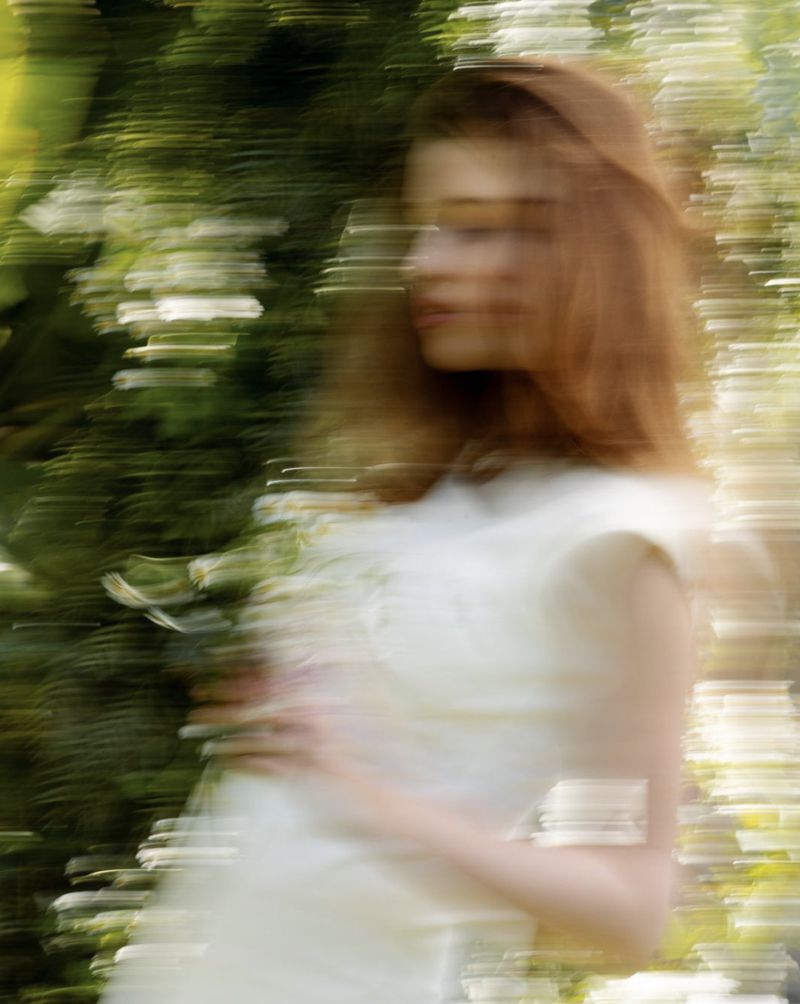 Dreamy, blurred image of an adult woman with long brown hair in a white dress, standing in a green outdoor garden setting with soft lighting.