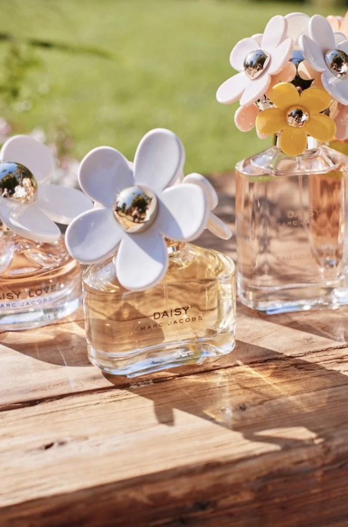 Three Marc Jacobs Daisy perfume bottles with distinctive flower caps resting on a rustic wooden surface outdoors.