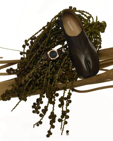 Studio shot of a black split-toe leather shoe, a gold wristwatch, and green berry branches on a white background, product photography.