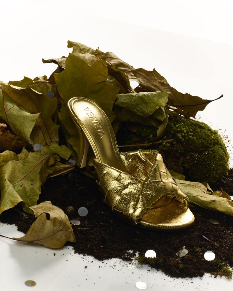 Studio shot of a gold Bottega Veneta mule-style high heel shoe resting on a bed of dry leaves, moss, and shiny confetti.