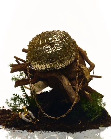 Golden chainmail clutch bag and necklace displayed on soil and moss with tree branches in a bright studio setup. 