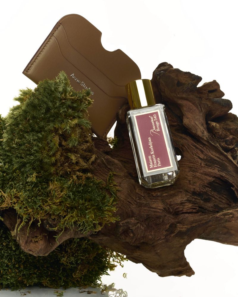 Close-up studio shot of Baccarat Rouge 540 perfume bottle with gold cap and a brown card holder on driftwood with green moss.