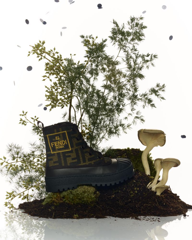 Fendi Roma boot on soil with green foliage and mushrooms against a white backdrop, accessorized with scattered black confetti.