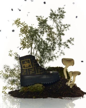 Fendi Roma boot on soil with green foliage and mushrooms against a white backdrop, accessorized with scattered black confetti.