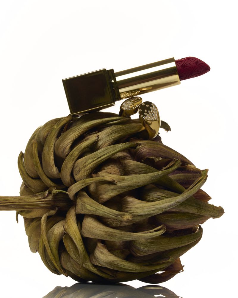Still life studio shot of an artichoke topped with lipstick and jewelry on a white background, showcasing color and texture.