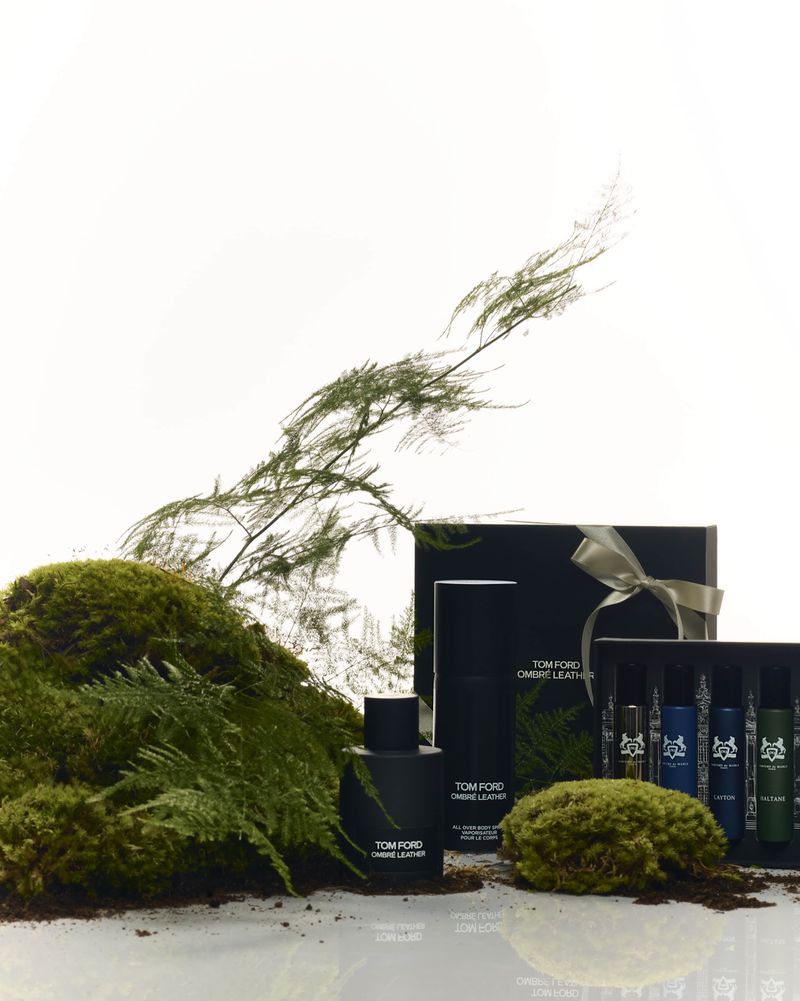 Tom Ford Ombre Leather products displayed with moss and greenery. Perfume bottle, gift box tied with ribbon, and smaller vials.