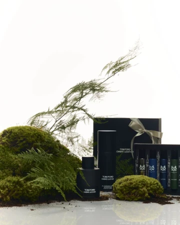 Tom Ford Ombre Leather products displayed with moss and greenery. Perfume bottle, gift box tied with ribbon, and smaller vials.