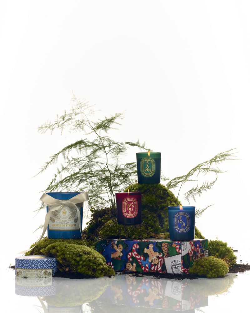 Aromatic candles in teal, red, and green glass jars are staged on moss and a colorful gift box, all against a white background.