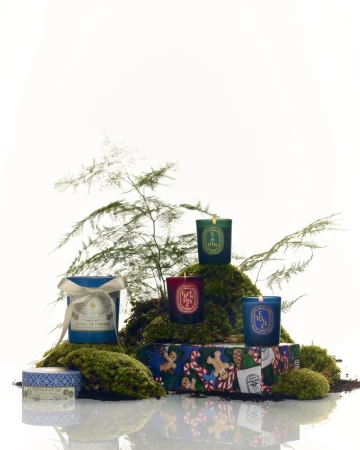 Aromatic candles in teal, red, and green glass jars are staged on moss and a colorful gift box, all against a white background.