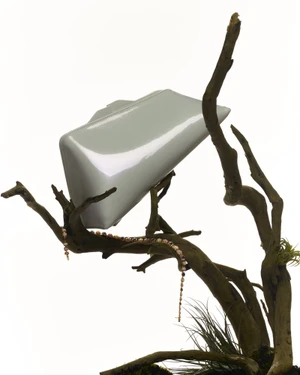 Glossy pale handbag resting on gnarled dark branches with draped beaded jewellery against a stark white background.