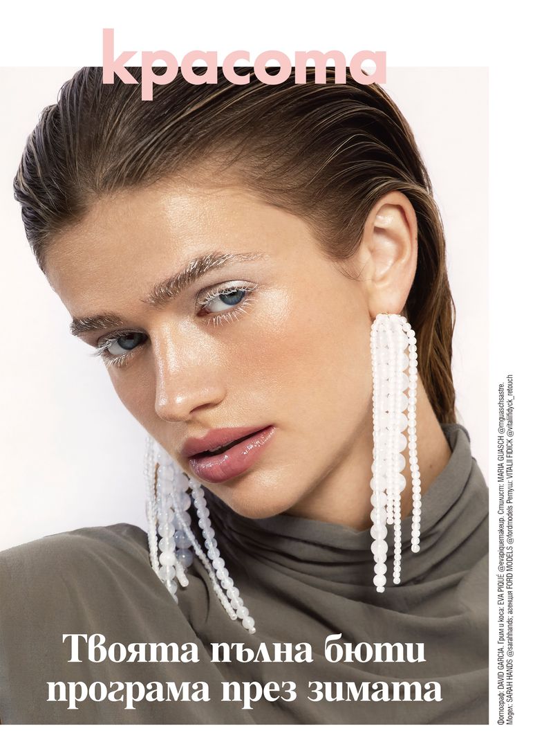 Close-up of a woman with slicked-back hair and long pearl earrings, wearing a muted-olive top, featured in a beauty editorial.