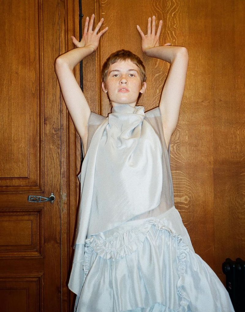 Woman in flowing light blue dress poses with hands above her head against a brown wood door indoors in a fashion style photograph.