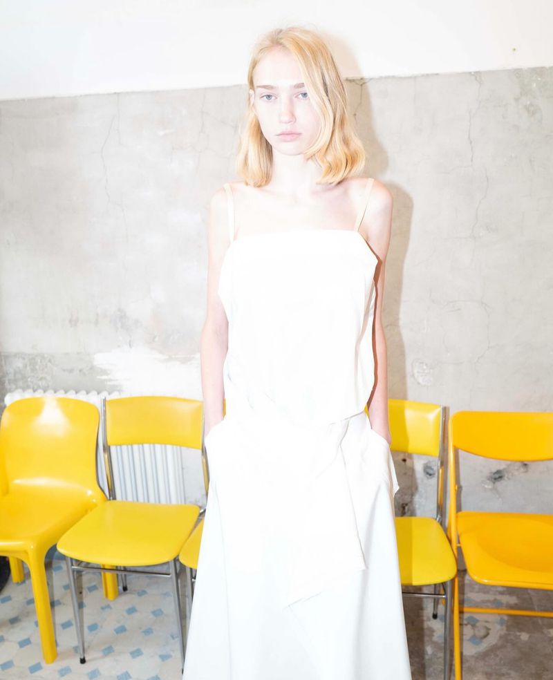 A fair-skinned young woman in white dress with a bare wall and yellow chairs, in a fashion or editorial setting under bright lighting.