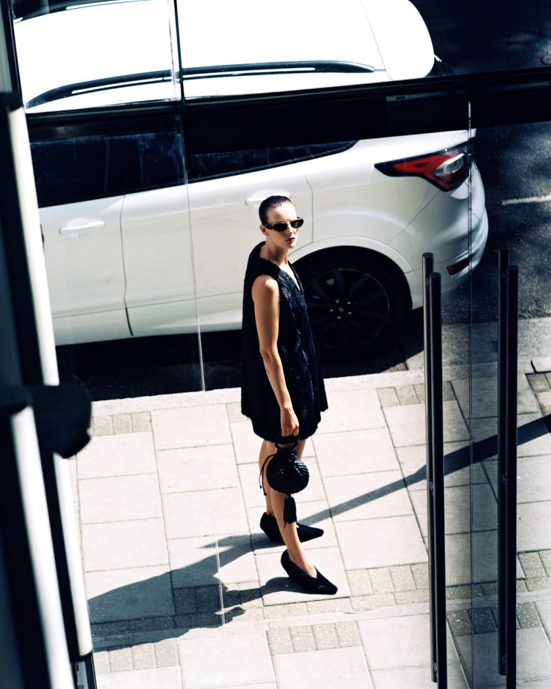 An adult woman standing outside next to a white SUV. She wears a black dress and sunglasses. The setting is an urban environment.