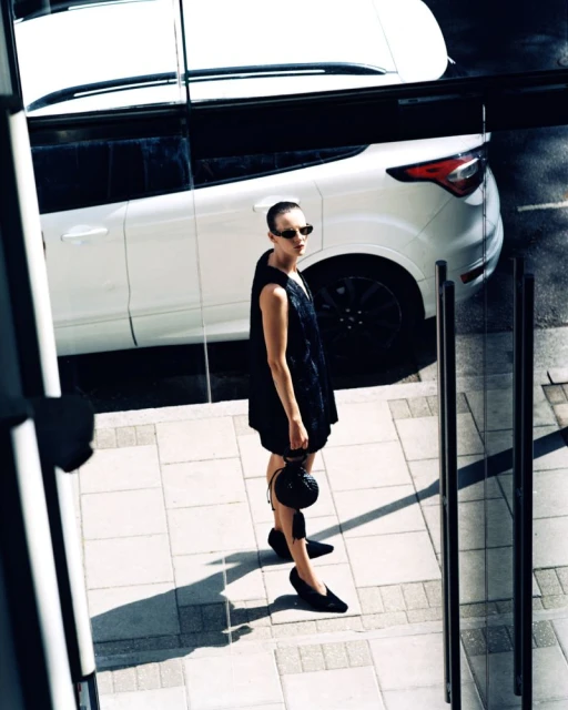 An adult woman standing outside next to a white SUV. She wears a black dress and sunglasses. The setting is an urban environment.