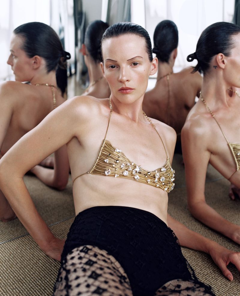 Fashion shot of a slender woman with slicked-back hair wearing a unique gold wire bra and black lace shorts, posing in an indoor setting.