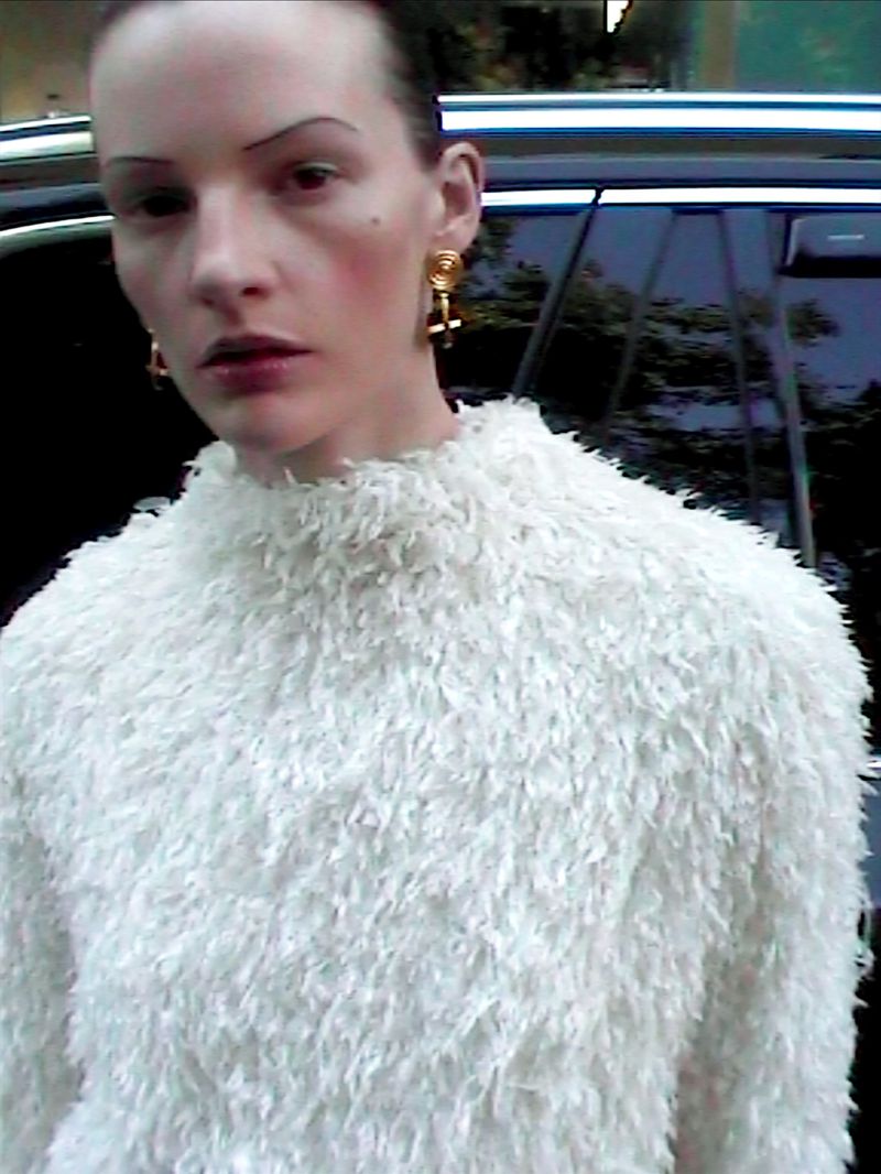 Close-up portrait of a slender adult woman with light skin wearing a white, fluffy sweater and gold earrings against a dark background.