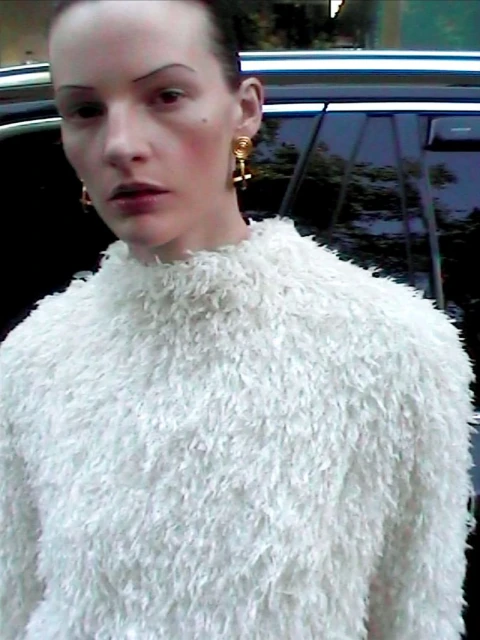 Close-up portrait of a slender adult woman with light skin wearing a white, fluffy sweater and gold earrings against a dark background.