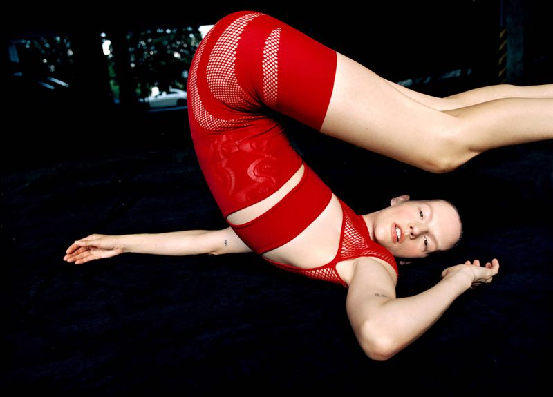 A light-skinned woman in red athletic wear lying on a dark surface with legs up in the air in a fitness or contortion pose.