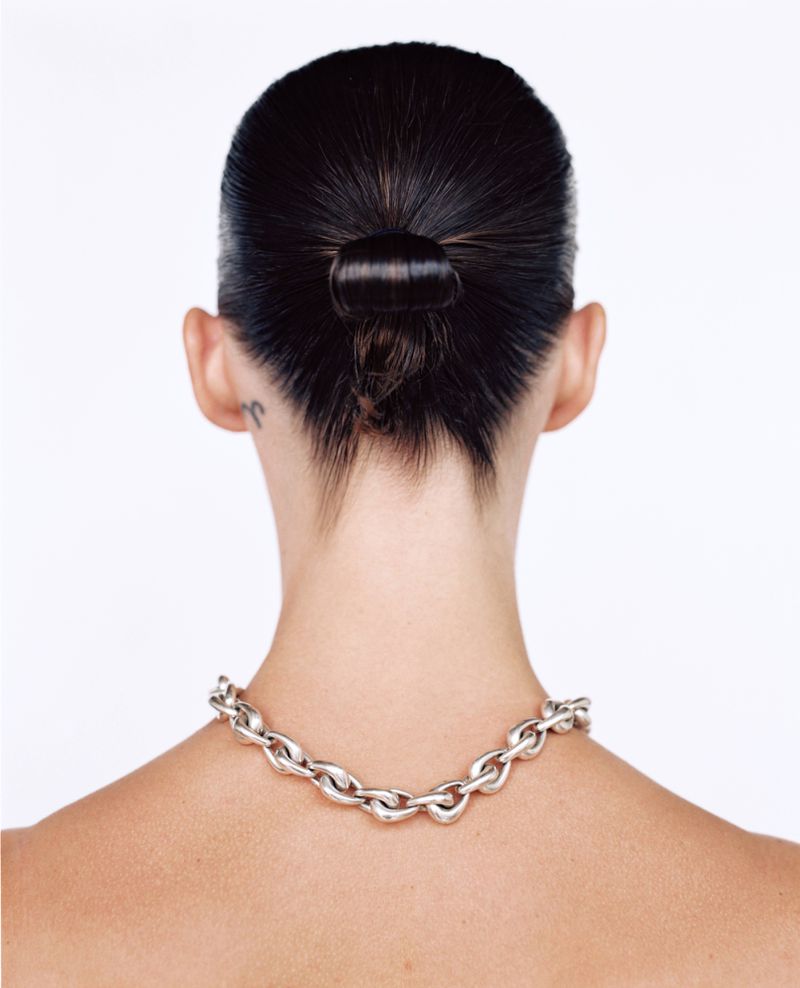 Back view of woman with dark hair in a bun wearing a silver chain necklace against a white backdrop in a studio setting.