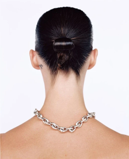 Back view of woman with dark hair in a bun wearing a silver chain necklace against a white backdrop in a studio setting.
