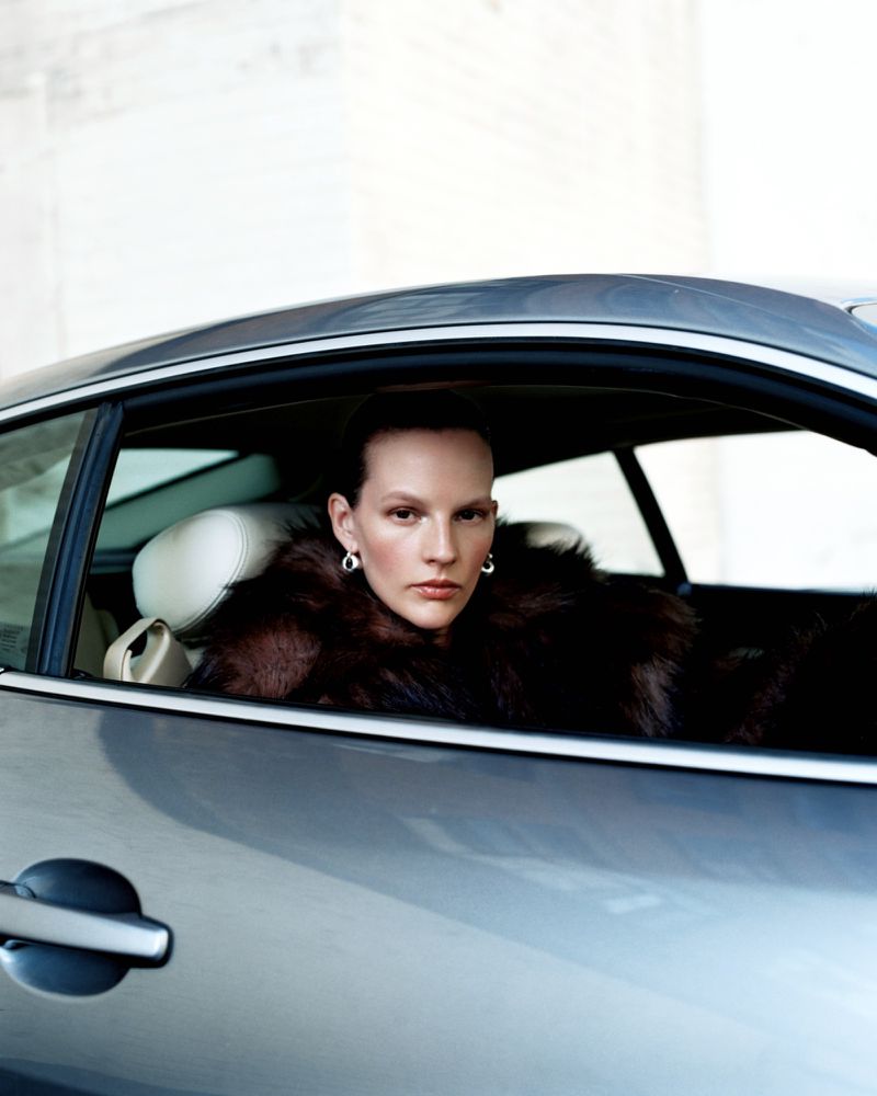 Woman in fur coat inside a silver car, looking at the camera with serious expression. Automotive and fashion themes, shot indoors.