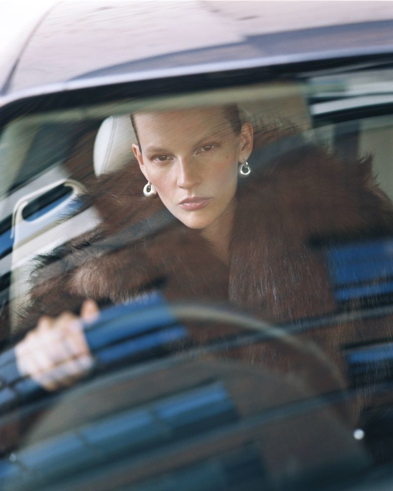 Woman with short hair and fur coat is driving a car, looking at the camera through the windshield. Fashion shoot.