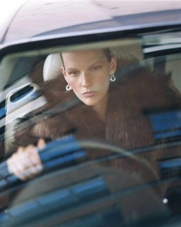 Woman with short hair and fur coat is driving a car, looking at the camera through the windshield. Fashion shoot.