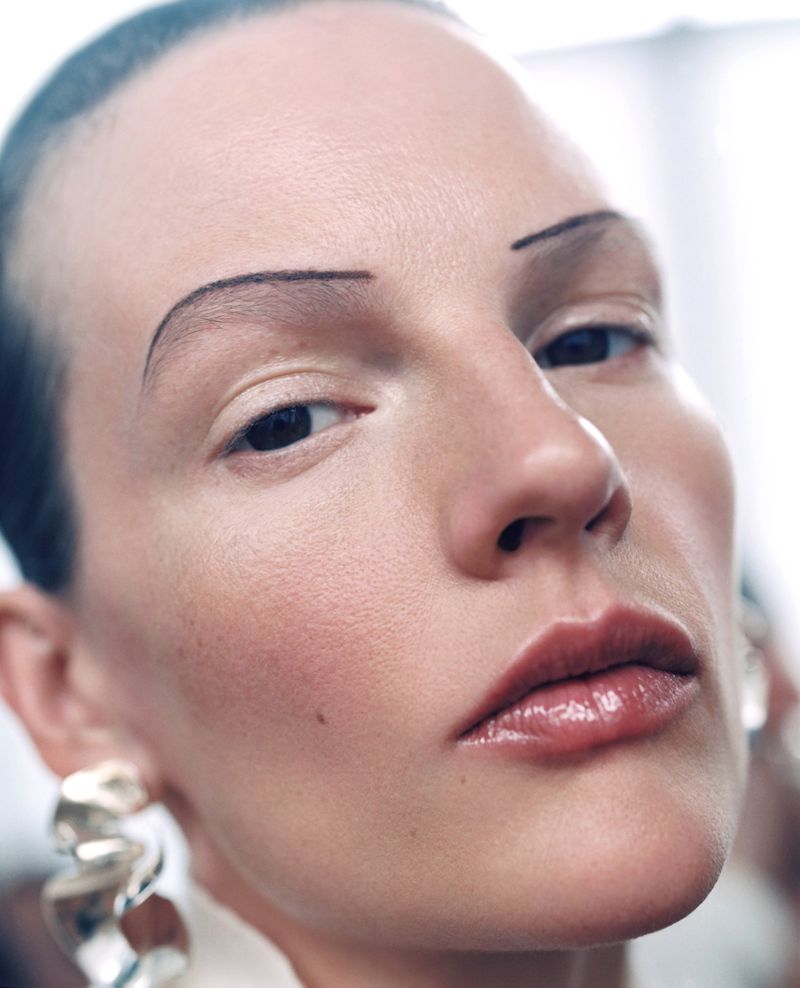Close-up portrait of a person with sleeked-back hair, distinctive eyebrows, glossy lips, and sculptural silver earrings in a neutral setting.