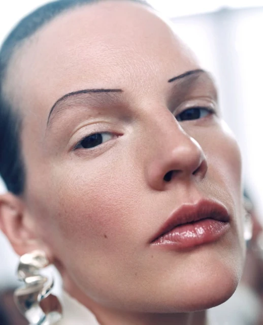 Close-up portrait of a person with sleeked-back hair, distinctive eyebrows, glossy lips, and sculptural silver earrings in a neutral setting.