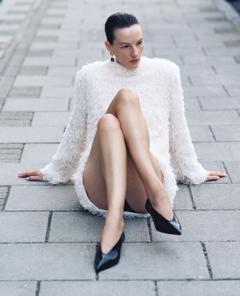 A woman in a white dress and black heels sits on a grey brick surface, looking away from the camera in an outdoor setting.