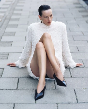A woman in a white dress and black heels sits on a grey brick surface, looking away from the camera in an outdoor setting.