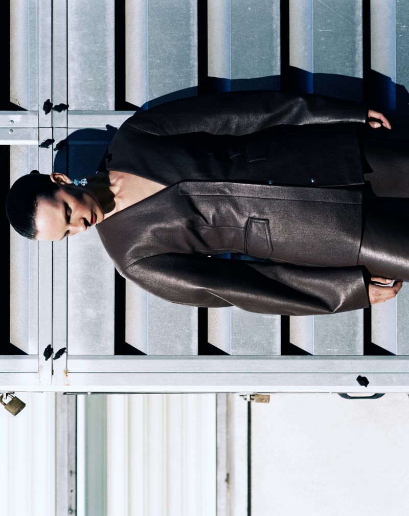 A woman poses in a brown leather jacket and pants in front of a metal grate and door on a sunny day, capturing a fashion-forward editorial shot.
