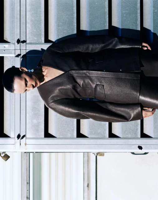 A woman poses in a brown leather jacket and pants in front of a metal grate and door on a sunny day, capturing a fashion-forward editorial shot.