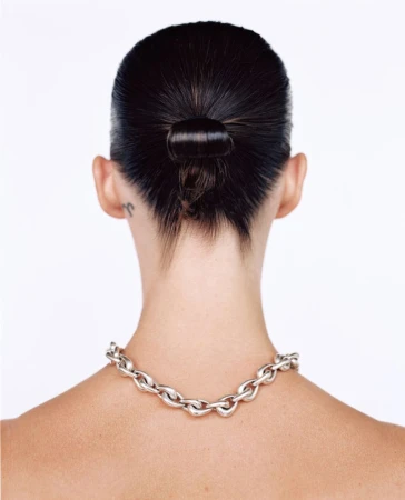 Rear view of a woman with slicked-back dark hair in a looped bun wearing a chunky silver chain necklace against a white background.