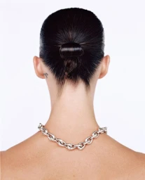 Rear view of a woman with slicked-back dark hair in a looped bun wearing a chunky silver chain necklace against a white background.