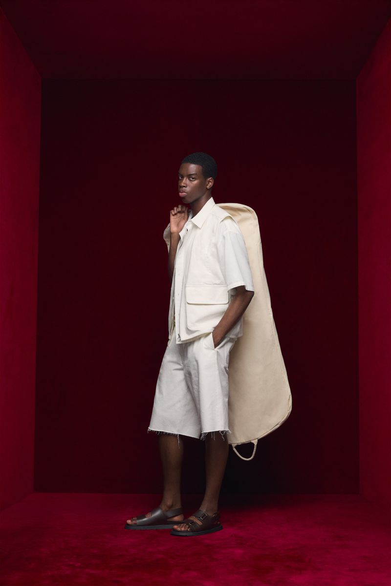 A Black male model poses in an indoor photo studio with a garment bag, wearing a light-colored short-sleeve shirt and shorts. The background is red.