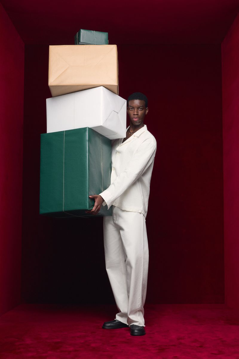 Adult man carries a large stack of wrapped presents indoors against a red wall and floor. He wears a white suit and black shoes.
