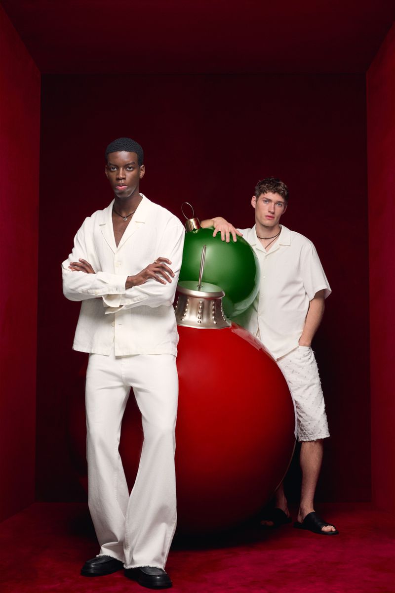 Two young men in white outfits pose with oversized red and green Christmas ornaments against a red studio backdrop, fashion editorial.