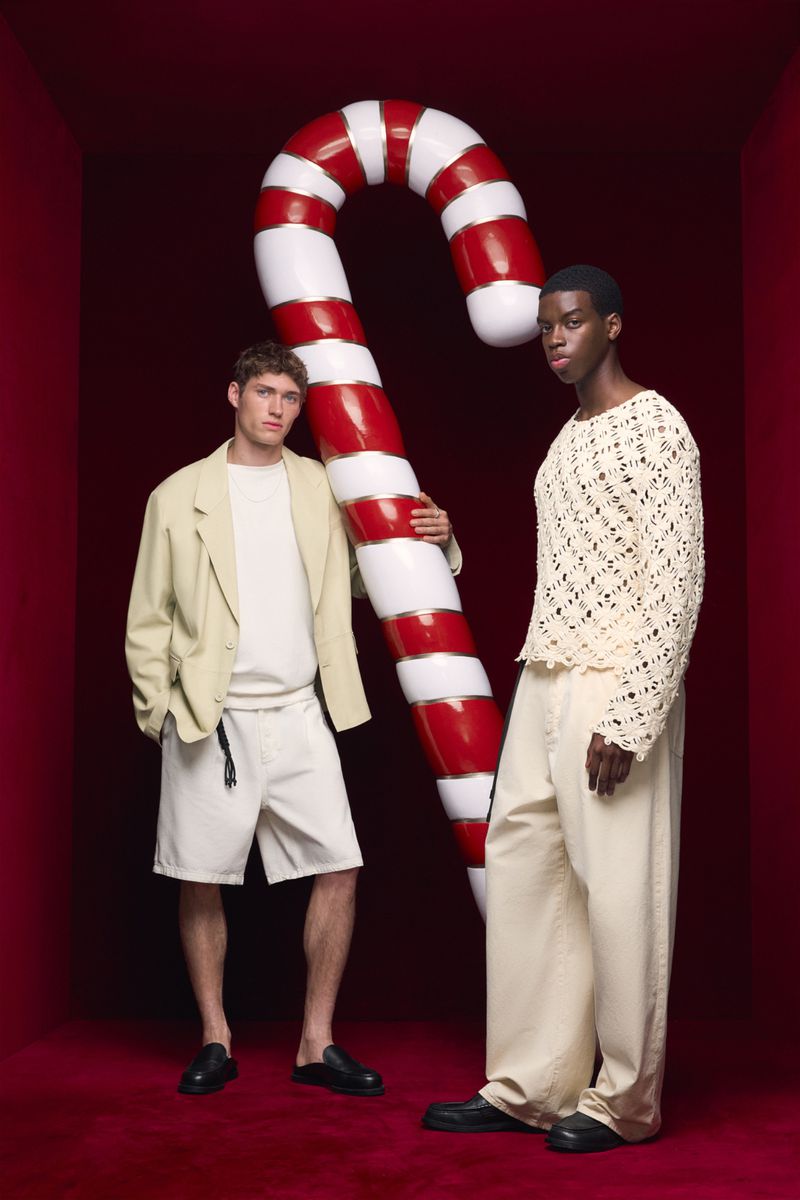 Two fashion models in beige tones pose in a studio setting with a giant candy cane, creating a visually striking, festive composition.