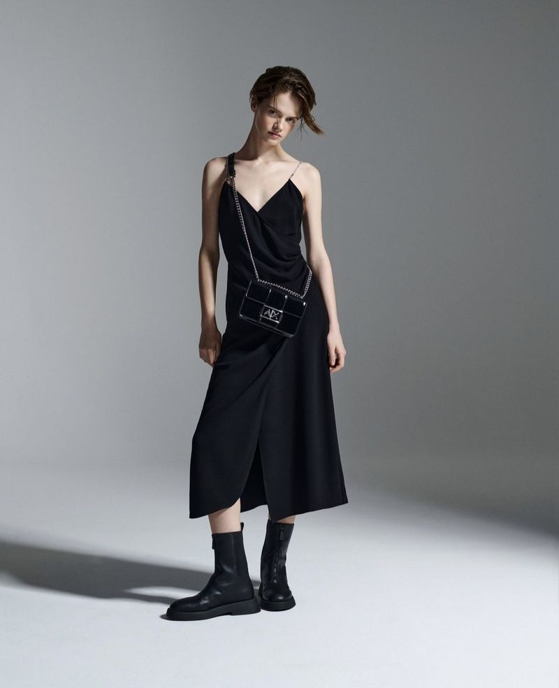 Woman poses in a black dress with a handbag in a studio, showcasing fashion and style against a neutral background, emphasizing elegance.