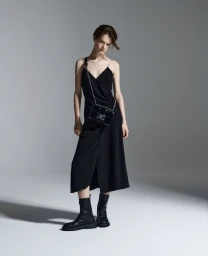 Model wearing a black slip dress and black boots holding a crossbody bag against a grey seamless studio background.